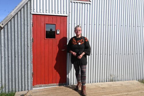 Kona í ullarpeysu, snýr fram á við, stendur á verönd fyrir framan silfurlitaða bylgjupappajárnsframhlið og vinstra megin við rauða hurð.